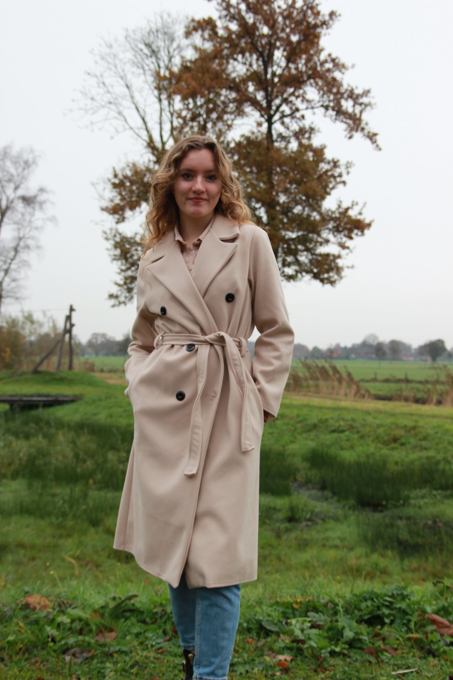 Beige trenchcoat met dubbele rij knopen en tailleriem, gestyled met ruffle blouse Saar