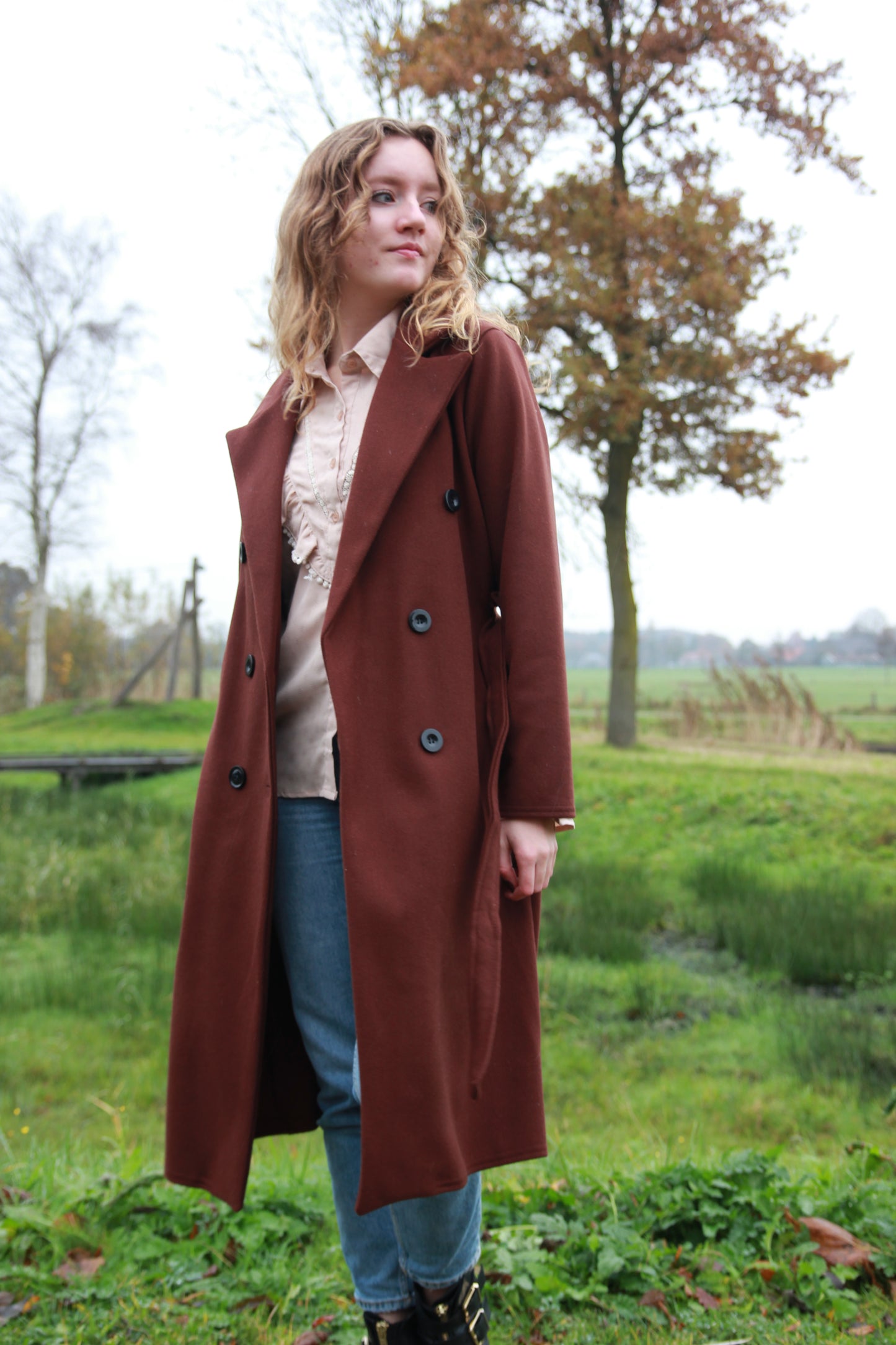 Donkerbruine trenchcoat met dubbele rij knopen en tailleriem, gestyled met ruffle blouse Saar